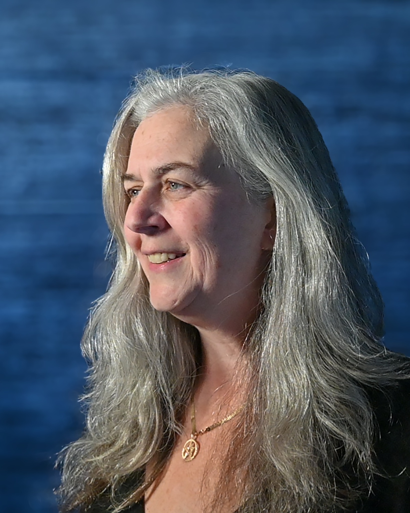 Portrait of Lissa Crosby, smiling warmly, with long silver hair, photographed against a deep blue background.